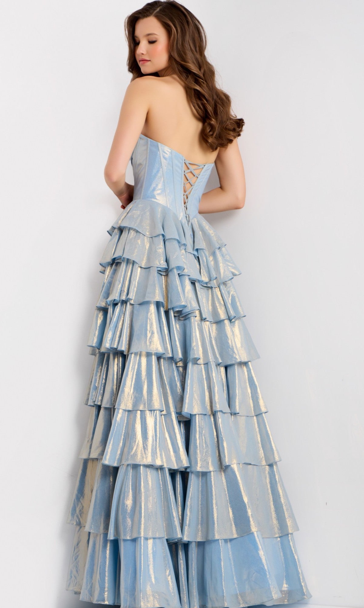 A woman stands against a white background wearing the Jovani 47030, a strapless long prom dress in light blue metallic fabric, featuring tiered layers and a lace-up corset bodice.